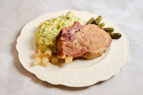 Roasted pork rib chop is placed on a plate next to creamy mashed potatoes and pickles. The dish is