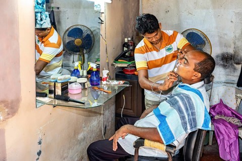 A hairdresser shaves a seated client in a simple room with a mirror and a fan, A man at a