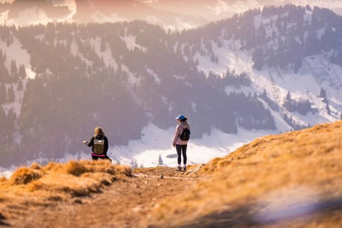Two people hiking a sunny mountainside with snow-covered valley in the background, Hochgrat