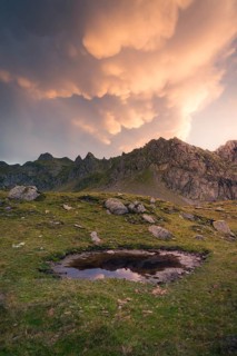 A tranquil mountain scene at sunset showcases dramatic, illuminated clouds above rugged peaks and