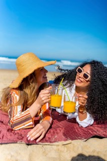 Two cheerful young women lying on a blanket on the sand, toasting with glasses of refreshing orange