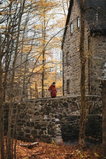 A woman walks across a stone bridge within a vibrant autumn forest in Quebec, Canada. The rustic