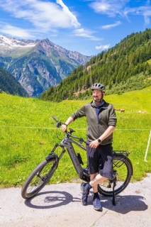 Man with helmet standing with his bicycle on a tarmac road, surrounded by green meadows and