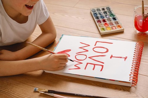 A young boy joyfully crafts a heartfelt Mother's Day card at home, using watercolor paint. The card