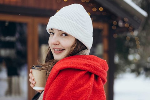 A young woman, wrapped in a bright red scarf, holds a coffee cup on a snowy winter day, smiling
