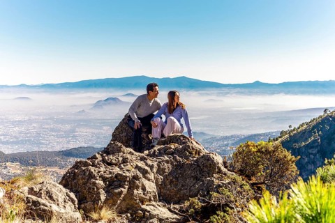 Two friends sitting on rocky terrain, surrounded by lush greenery, admire a breathtaking mountain