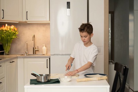 A young boy focuses on preparing a meal in a modern kitchen. He uses a knife on a white counter