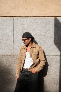A man stands casually in front of a stone wall, wearing sunglasses, a flat cap, a beige jacket, and