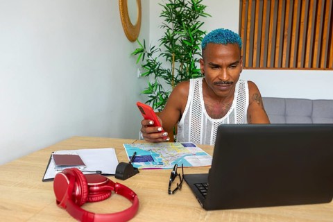 A blue-haired LGBTQIA+ man plans a holiday trip with a laptop and phone. Maps, headphones, and