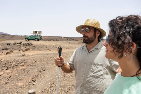 A blind man with a cane and a woman stand together in a desert landscape under the bright sun,