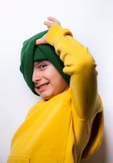 A playful child wearing a bright yellow sweatshirt and green beanie strikes a cheerful pose. The