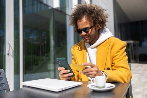 Afro man in a stylish yellow coat and sunglasses is sitting at an outdoor cafe table, smiling while