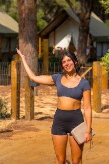 Young tourist woman with a clutch bag waving hand and smiling at the entrance of a glamping resort,