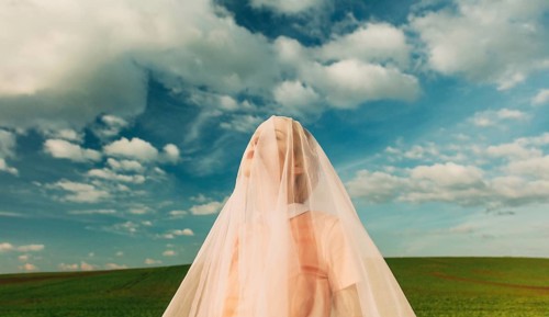 A woman stands outdoors, draped in sheer fabric, with a serene expression. The backdrop is a lush