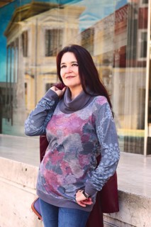 A woman stands confidently outdoors, wearing a stylish floral sweater and jeans. The reflection of