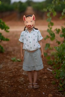 A child stands on a dirt path in a playful pig mask, surrounded by lush greenery and vineyards.