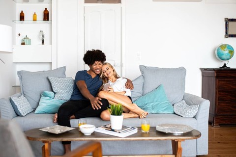 A happy couple sits on a cozy couch, looking at a phone. The bright living room has modern decor,