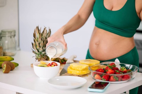 A pregnant woman in a green yoga outfit prepares a healthy breakfast with fresh fruits and milk.