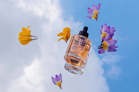 A glass serum skincare bottle with golden liquid is surrounded by vibrant yellow and purple flowers