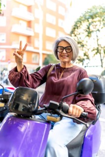 Senior woman with grey hair and eyeglasses smiling, riding a purple scooter through an urban