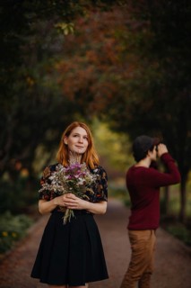 A woman holding a bouquet of wildflowers stands thoughtfully under colorful autumn trees, while a