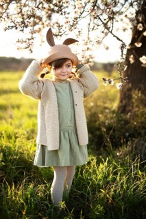 A young girl dressed in a green dress and a bunny ears hat stands amidst blooming almond blossoms,