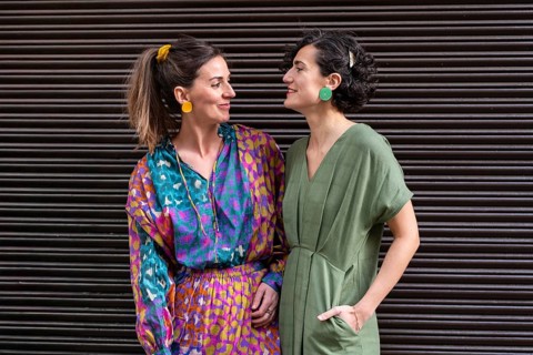 Two stylish women stand in cheerful clothing, gazing at each other with smiles. They are wearing
