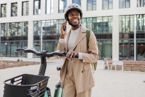 A fashionable black woman in an urban setting stands with a bike, wearing a helmet and talking on