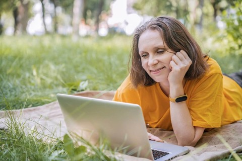 A woman is lying on a blanket outdoors in a park, using her laptop for work. She enjoys the serene