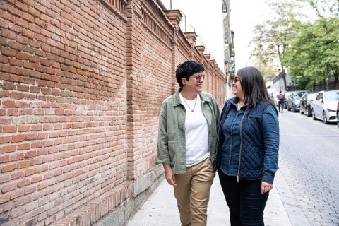 A curvy lesbian couple walks happily along a brick wall, sharing a joyful moment on a sunny day.