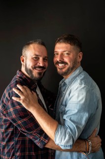 A joyful gay couple embraces with smiles, dressed in casual plaid and denim shirts against a dark