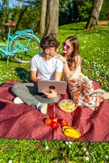 Romantic couple having picnic in park, watching movie on laptop, surrounded by food and drinks,