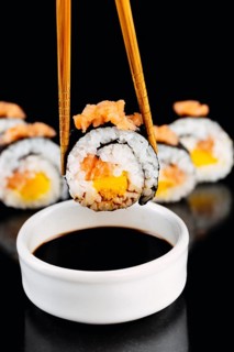 Close up of a sushi roll with salmon and mango, held by bamboo chopsticks above a bowl of soy sauce