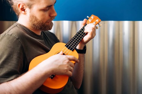 A musician with a beard passionately plays a bright orange Mexican ukulele, focused on his music.