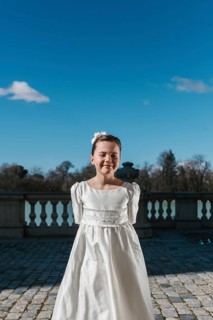 A young girl stands confidently in her white First Communion dress, under a clear blue sky. The