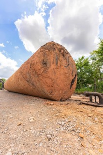 A large, brown tank lies flat on a construction site, surrounded by sky, clouds and trees,