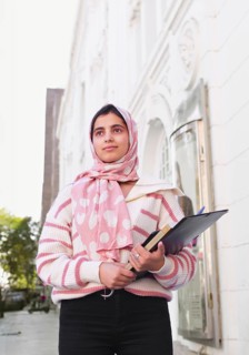 A arab student woman wearing a pink hijab stands outdoors, holding books and a clipboard. She is