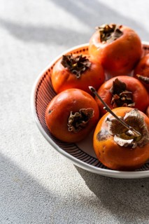 A bowl filled with freshly picked, ripe persimmons, positioned on a light textile surface, bathing