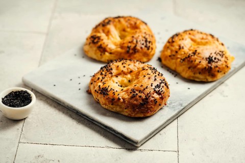 Three golden-brown bagels sprinkled with black sesame seeds rest on a marble board. A small bowl of