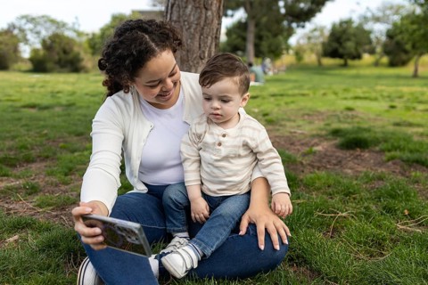 A mother and her baby share a joyful moment in the park. They sit together on the grass, surrounded