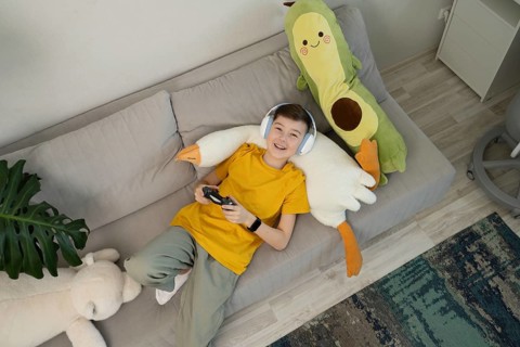 A happy boy wearing headphones plays video games while lounging on a cozy sofa. Surrounded by plush
