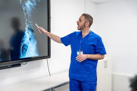 Medical professional in scrubs pointing to a large screen displaying an x ray of the human spine