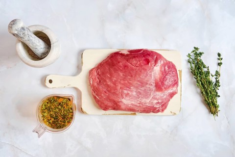 Pork shoulder on a cutting board ready for cooking. Fresh thyme and a bowl with spices are nearby.