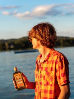 Young man standing by the lake, holding a bottle of alcohol in his hands, herbal liqueur, red hair,