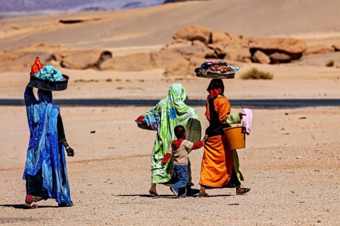 Group of people in traditional clothing carrying loads on their heads in a desert landscape, woman