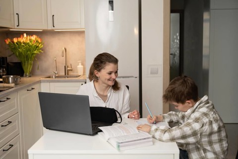 A mother helps her son with homework at the kitchen table. A laptop and books are present,
