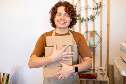 A cheerful worker in a shop setting holds cardboard boxes, wearing a brown shirt and apron A