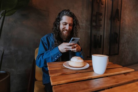A man with long hair and a beard smiles while using his smartphone at a wooden table in a cozy cafe