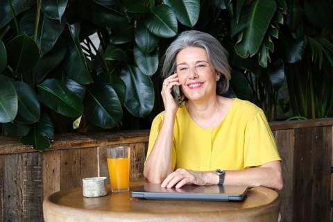 Mature woman, smiling in a yellow blouse, enjoys a phone call while sitting at a wooden table with
