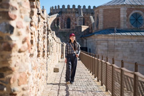 An Asian woman in casual attire walks along the cobbled medieval walls of Avila, Spain, surrounded